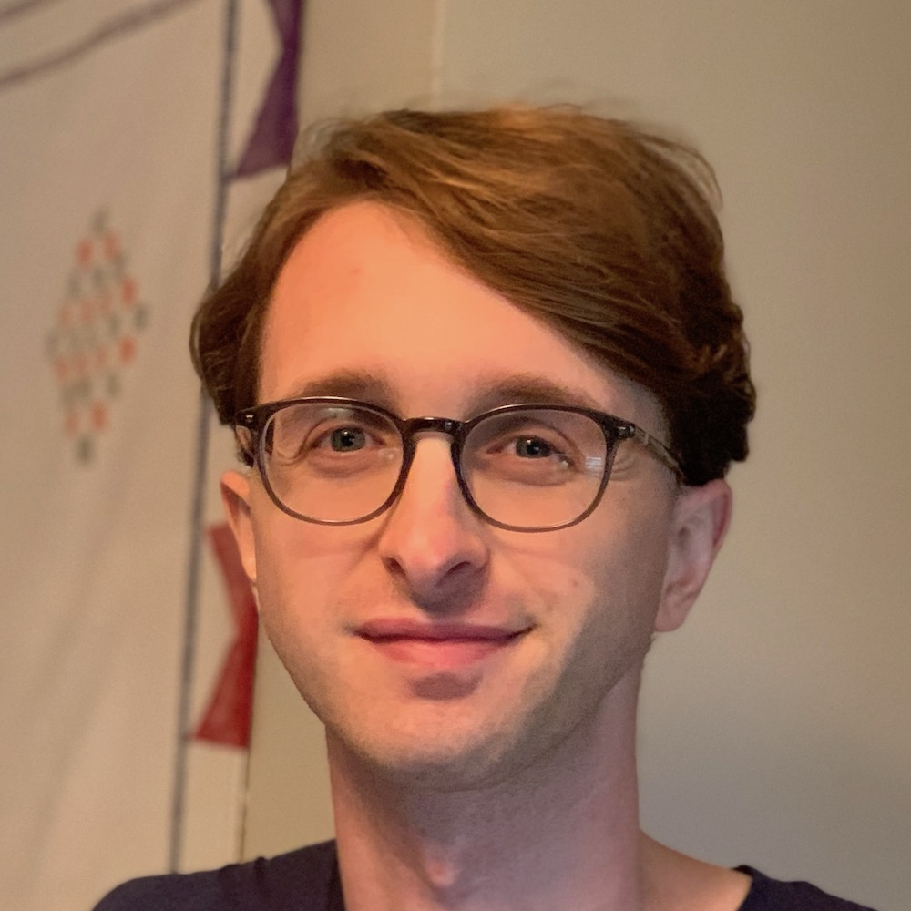 Evan Minto looking up and slightly smiling. He’s a young white man with medium-length hair and black plastic glasses.